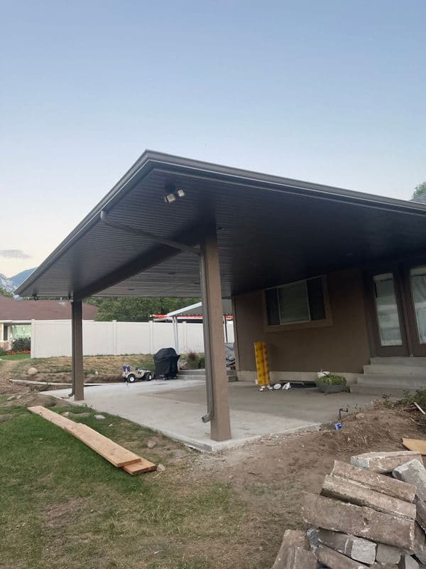 Patio cover with metal roof and support beams, featuring a concrete slab and garden area.