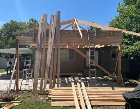 Home construction in progress with wooden beams, ladders, and lumber in a sunny backyard.